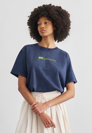 Kobieta z afro fryzurą, ubrana w granatowy T-shirt Nike Athletics Club oraz jasnobeżową plisowaną spódnicę na jednolitym tle.
