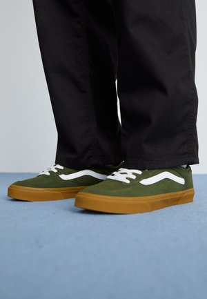 Grønne semskede sneakers med hvide striber og brune gummisåler, snøret med hvide snørebånd, parret med mørke bukser mod en blå baggrund.