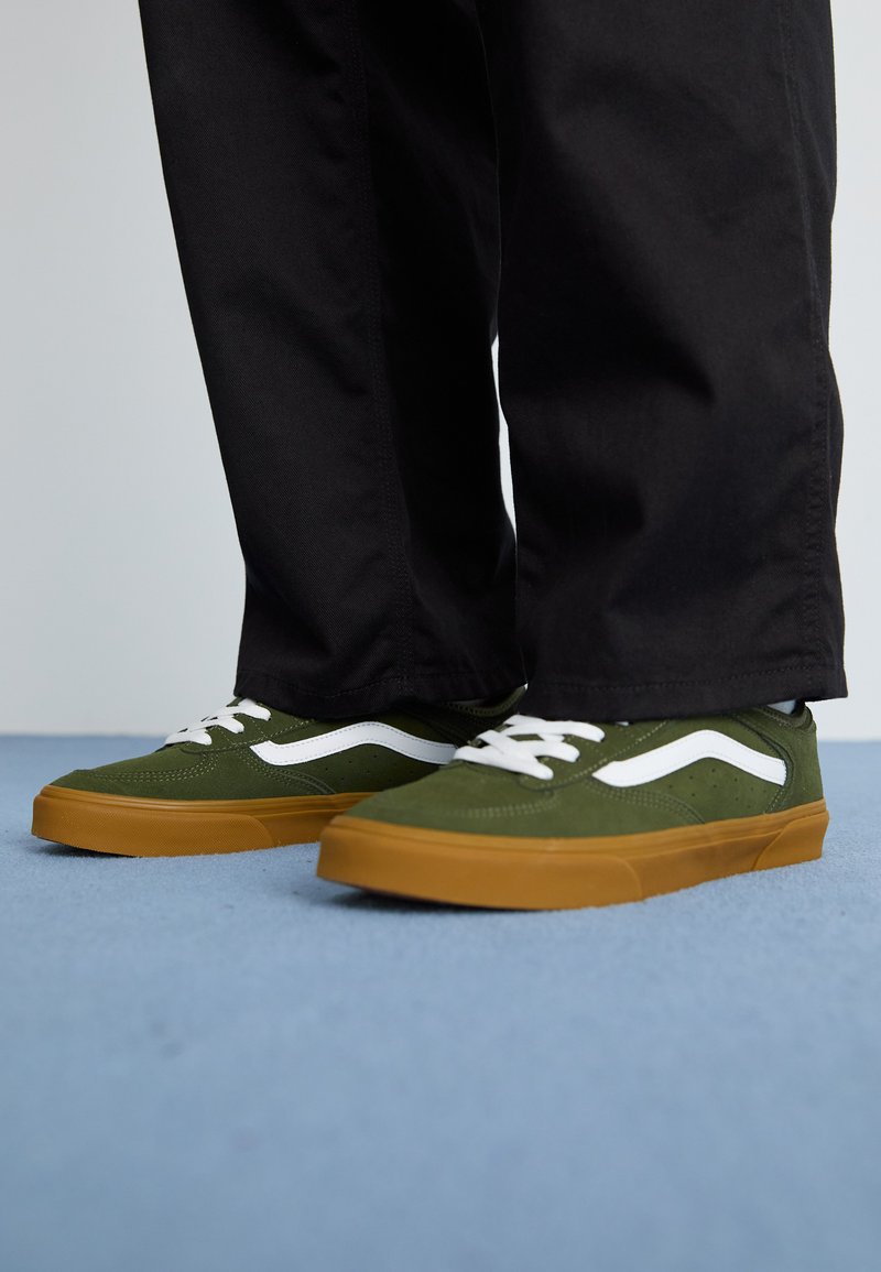 Grønne ruskindsneakers med hvide striber og brune gummisåler, snøret med hvide snørebånd, iført mørke bukser foran en blå baggrund.