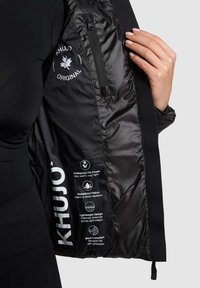 Musta takki, jossa sileä ja vedenpitävä vuori. Takissa on vetoketjullinen tasku ja suuri valkoinen logo. Hoito-ohjeissa korostetaan eristystä ja tuulenpitävyyttä.