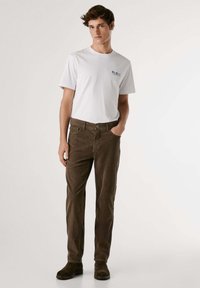 Camiseta blanca de cuello redondo con un pequeño logo azul, combinada con pantalones de pana marrones. El modelo posa con las manos en los bolsillos, llevando botas de tobillo marrones.