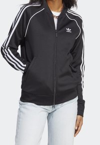 Person som bär en svart Adidas zip-up jacka med vita ränder på ärmarna och logotyp, tillsammans med ljusblå jeans och ett kedjehalsband.