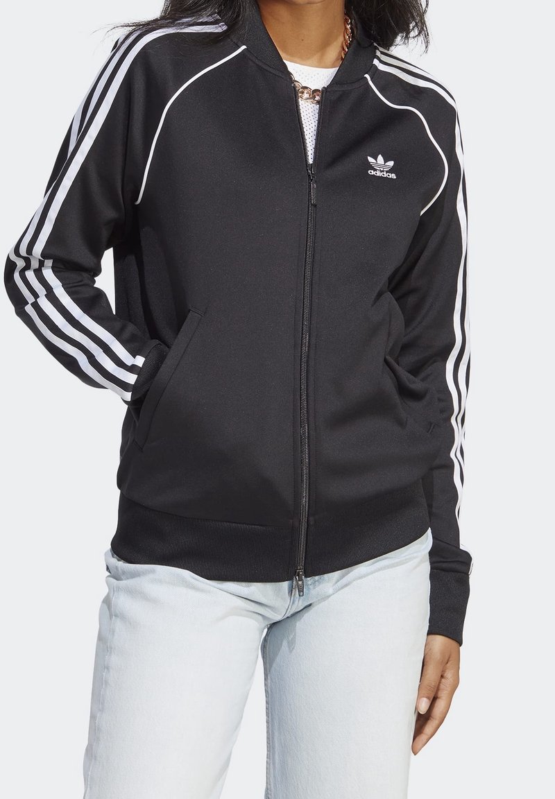 Person som bär en svart Adidas zip-up jacka med vita ränder på ärmarna och logotyp, tillsammans med ljusblå jeans och ett kedjehalsband.