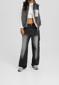 Szary polarowy kamizelka z zamkiem z przodu, biały krótki top i czarne dżinsy o szerokich nogawkach z wyblakłym wzorem i przednią kieszenią. Szare sneakersy dopełniają całość.