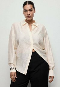 Woman with dark hair in a low bun wearing a sheer, cream button-up blouse and black pants, standing against a plain light background.
