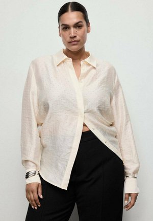 Mujer con cabello oscuro en un moño bajo, vestida con una blusa de botones crema y transparente y pantalones negros, de pie contra un fondo claro y liso.