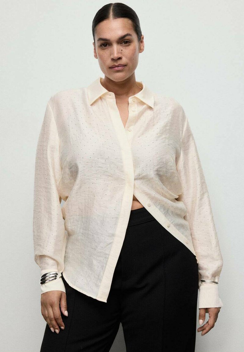 Woman with dark hair in a low bun wearing a sheer, cream button-up blouse and black pants, standing against a plain light background.