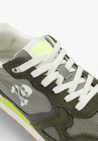 Baskets grises et olive avec lacets blancs, logo tête de mort et os croisés sur le côté, accents jaune néon et panneaux en tissu texturé.