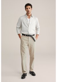 Mann trägt hell gestreiftes Hemd über weißem T-Shirt, beige Hose mit schwarzem geflochtenem Gürtel und schwarzen Loafers, steht mit den Händen in den Taschen.