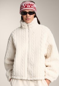 Jachetă fleece de culoarea crem, cu guler �înalt, model texturat cu torsadă și închidere cu fermoar. Asortată cu un bandană roșie și albă cu imprimeu grafic și ochelari de soare.