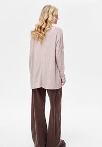 Jasnoróżowy, długi sweter z długim rękawem o luźnym kroju i zaokrąglonym podłożu; w połączeniu z szerokimi brązowymi spodniami, tworzącymi miękki, casualowy zestaw.