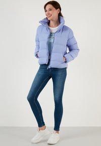 Šviesiai violetinė pūkų striukė su aukštu apykakle ir užtrauktuku. Derinama su mėlynomis siaurėjančiomis džinsinėmis kelnėmis ir baltais sportbačiais, prieš neutralų foną.