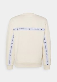 Sweat-shirt crème à manches longues avec des bandes blanches arborant les logos "Calvin Klein Jeans" et "CK" au dos et le long des manches.