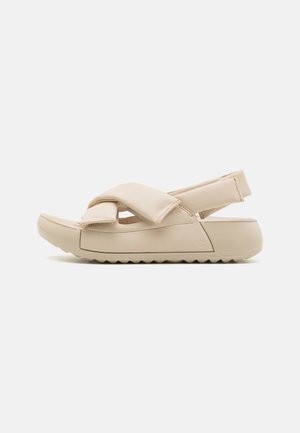 Sandalo beige con cinturini incrociati, materiale superiore testurizzato e suola spessa e rigata. Il design presenta un retro aperto per un facile indossamento.