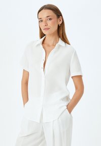 Camicia bianca a maniche corte con colletto, caratterizzata da chiusura frontale con bottoni e un taglio morbido, realizzata in tessuto leggero.
