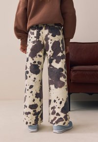 Jean à jambes larges à imprimé vache en crème et marron foncé, avec une taille haute, des poches arrière et une texture denim douce, associé à des baskets bleu clair.