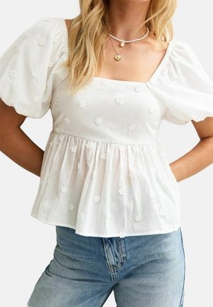 Frau trägt weiße Bluse mit Puffärmeln und quadratischem Ausschnitt sowie bestickten Blumendetails, kombiniert mit hellblauen Jeans.