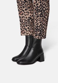 Black leather ankle boots with a square toe, low block heel, and smooth texture. Worn with leopard-print patterned trousers.