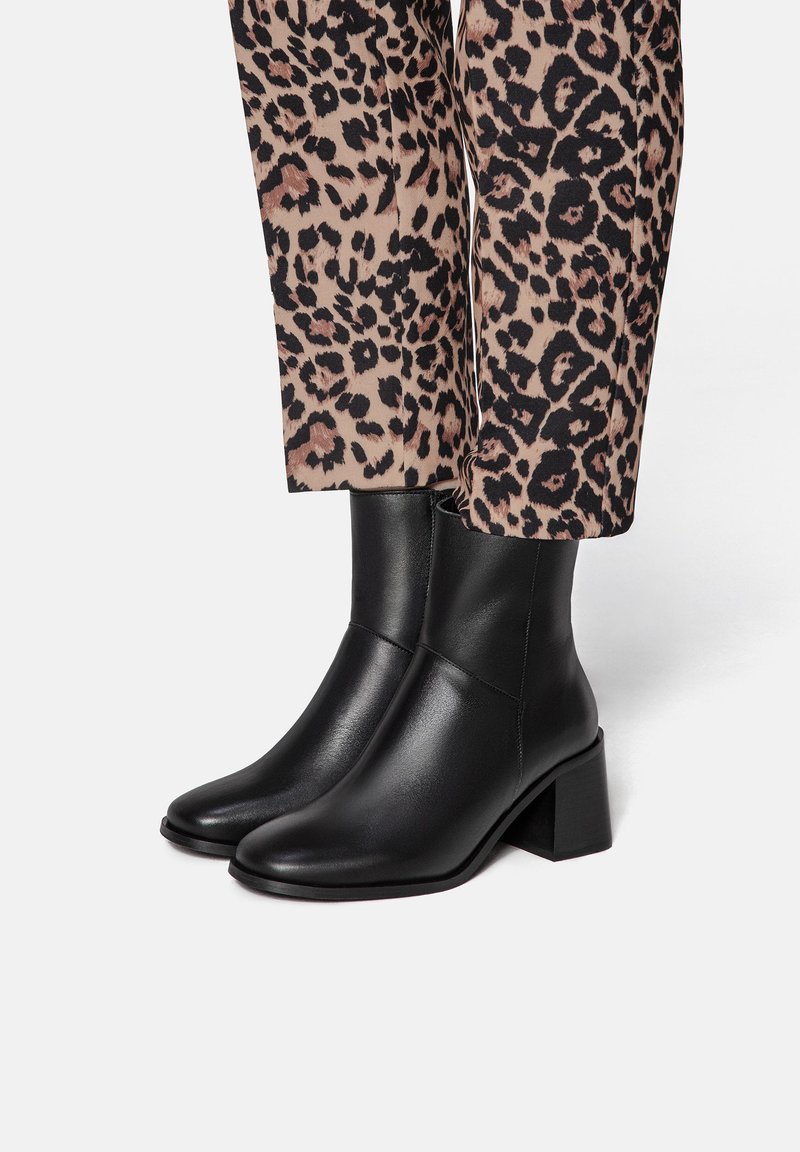 Black leather ankle boots with a square toe, low block heel, and smooth texture. Worn with leopard-print patterned trousers.