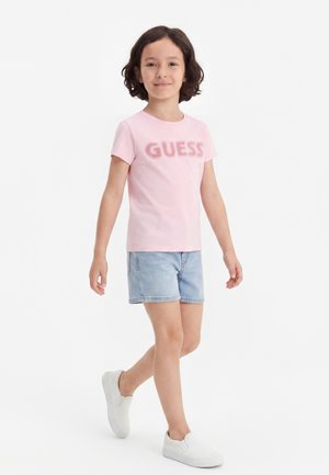 Lille barn gående, iført en lyserød "GUESS" t-shirt, lyse denimshorts og hvide slip-on sko mod en ensfarvet hvid baggrund.