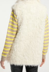 Biała kamizelka z futra syntetycznego o teksturowanej powierzchni, nałożona na sweter w paski z poziomymi pasami w odcieniach szarości, żółci i beżu.