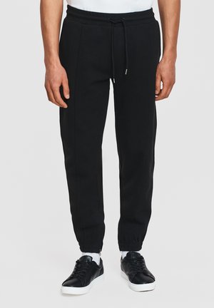 Mężczyzna noszący czarne spodnie dresowe z wiązaniem w pasie i elastycznymi ściągaczami, w połączeniu z czarnymi trampkami i białymi skarpetkami.