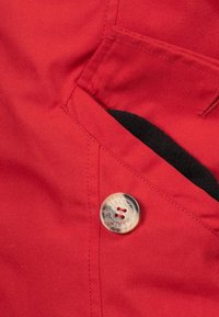 Tessuto rosso con una superficie liscia, presenta una patta per la tasca e un bottone rotondo lucido con due fori. L'interno è foderato di nero.