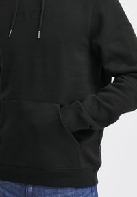 Black pullover hoodie made of soft fabric. Features a front pocket, ribbed cuffs, adjustable drawstrings, and "TOKYO" text embossed on the chest.