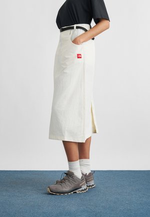 Osoba ubrana w białą spódnicę midi z bocznym rozcięciem, czarny pasek, czarny top, białe skarpetki i szare buty sportowe Salomon stoi na niebieskim dywanie.