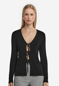 Svart, glittrande kavaj med djup V-ringning och knytning fram. Långa ärmar och avslappnad passform. Framhävd av en subtil glittrande textur.