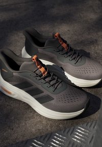 Paar graue und schwarze Laufschuhe mit orangen Zuglaschen und weißen Sohlen auf strukturierter Bodenoberfläche.