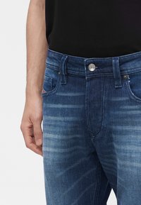 Homme portant un jean bleu foncé délavé et une chemise noire, avec la main gauche reposant près de la poche sur un fond clair uni.