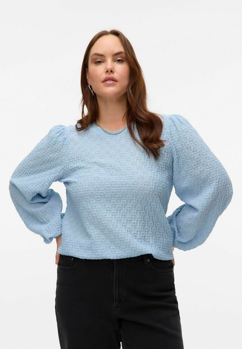 Blouse texturée bleu clair avec des manches longues bouffantes et un col côtelé, associée à un pantalon noir.
