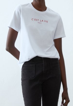 Person iført en hvid T-shirt med rød tekst "C'EST LA VIE PARIS" og sorte jeans, stående mod en ensfarvet lys baggrund.