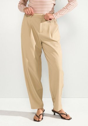 Femme portant un pantalon beige taille haute à jambes larges et des sandales noires à talons, ajustant la taille, avec un haut à manches longues rose et blanc rayé.