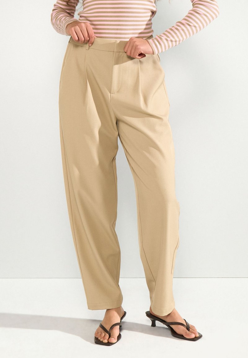 Femme portant un pantalon beige taille haute à jambes larges et des sandales noires à talons, ajustant la taille, avec un haut à manches longues rose et blanc rayé.