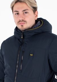 Granatowa parka z wysokim kołnierzem i kapturem, z przednim zamkiem, kieszenią na piersi oraz żółtymi akcentami stębnowania. Gładka faktura materiału.