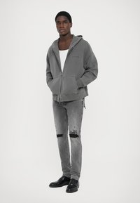 Grå zip-hoodie med en avslappnad passform, som har en stor ficka och en robust textur, ihop med slitna grå jeans och svarta skor.