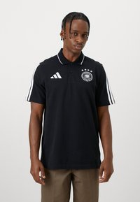 Muž v černém polokošili Adidas s bílými pruhy na rukávech a logem Deutscher Fussball-Bund, stojící na jednobarevném bílém pozadí.