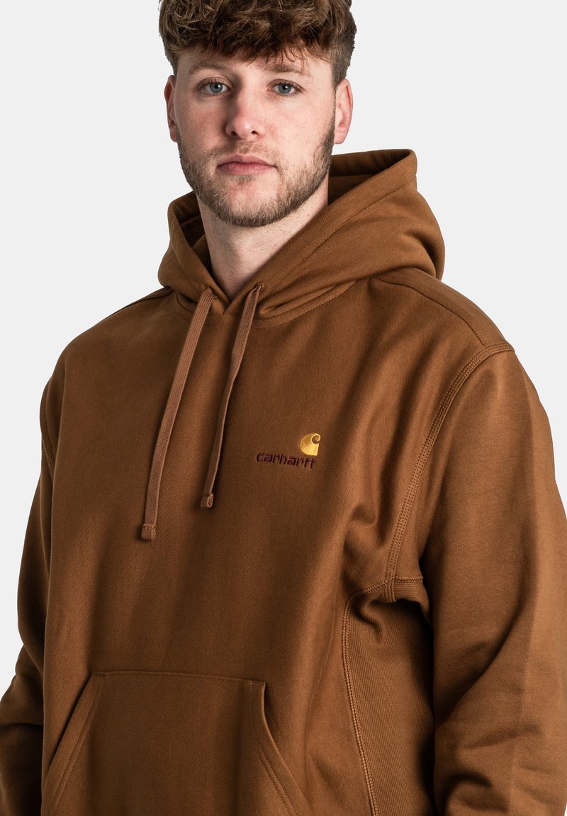 Sudadera marrón con bolsillo frontal, capucha ajustable con cordón y logotipo bordado en dorado y burdeos en el pecho. Textura de tela suave.