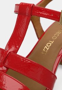 Sandale en cuir verni rouge avec plusieurs brides croisées, une texture lisse et une semelle intérieure marron ornée d'une marque dorée.