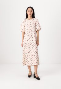 Witte jurk met rood bloemenpatroon, korte pofmouwen en een getailleerde taille. Het model draagt zwarte lakleren Mary Jane schoenen. Vloer lengte silhouet.