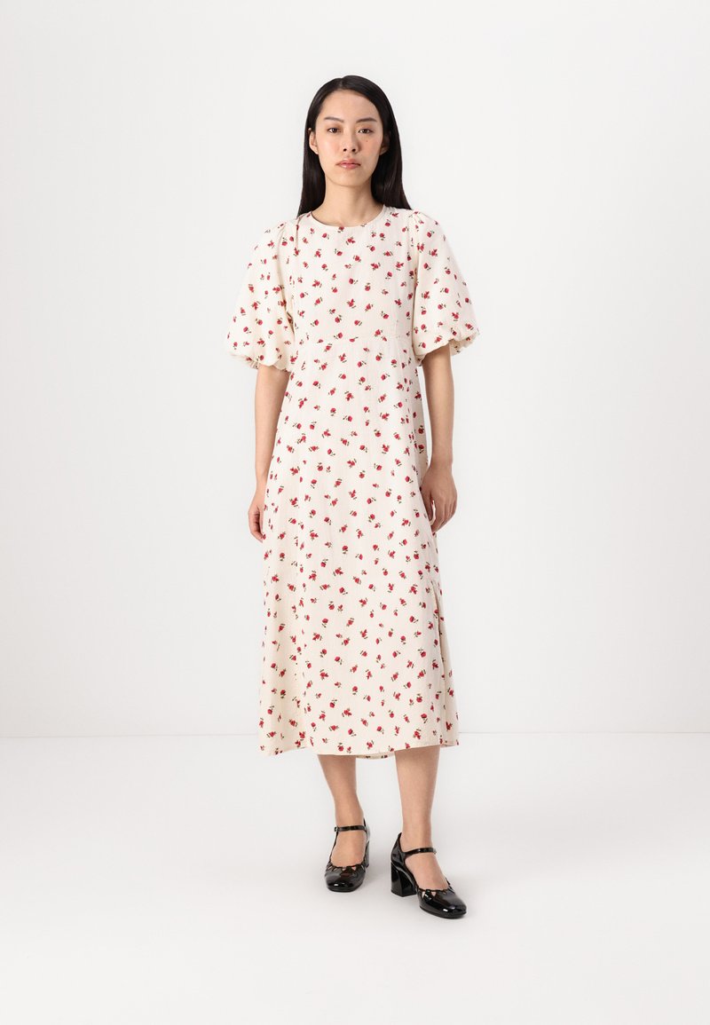Witte jurk met rood bloemenpatroon, korte pofmouwen en een getailleerde taille. Het model draagt zwarte lakleren Mary Jane schoenen. Vloer lengte silhouet.