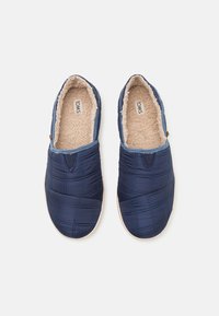 Navy blue quilted slip-on shoes with a soft beige fur lining and padded texture. Features stitched accents and light-colored soles.