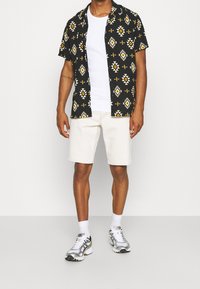 Svart t-shirt med kort ärm och geometriskt mönster i gult och vitt, tillsammans med ljusbeiga shorts och silverfärgade träningsskor.