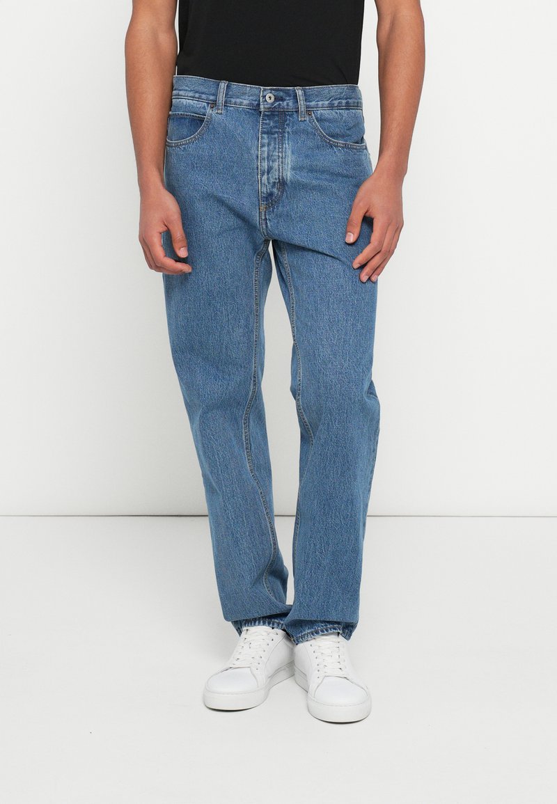 Vaqueros de denim azul claro con un diseño de pierna recta, cinco bolsillos y cierre de botón. Combinados con una camiseta negra y zapatillas blancas.