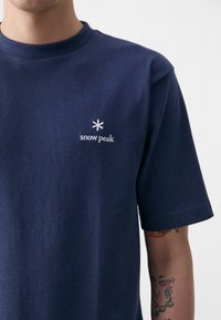 Tamsiai mėlynas medvilninis marškinėliai su apvaliu kaklu. Ant kairės krūtinės dalies yra mažas baltas išsiuvinėtas "sniego viršūnė" logotipas. Trumpomis rankovėmis.