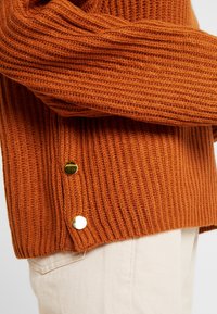 Orange ribbstickad tröja med två guldfärgade knappar på sidan, bärs över ljusbeige byxor.