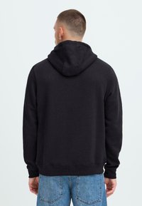 Hombre con cabello corto y arete de aro, vistiendo una sudadera negra y jeans azules, mostrado de espaldas contra un fondo claro.