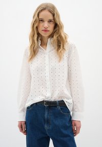 Camicia bianca con bottoni a quadri e piccoli ritagli, abbinata a jeans in denim blu a vita alta e una cintura nera.